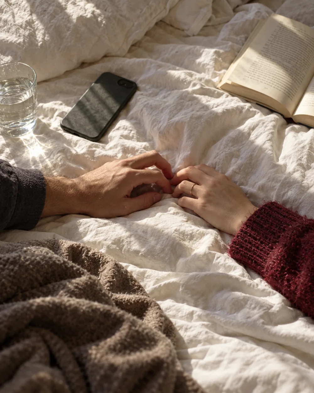 Deux mains qui se frôlent sur des draps, un téléphone posé face contre le lit